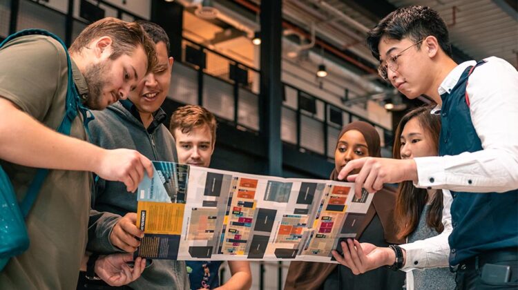 Six students are standing and gathered around a leaflet together