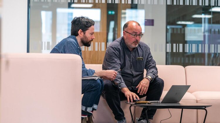 Image of two members of staff sitting on sofas in MECD looking at a laptop screen together