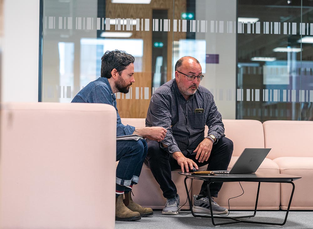 Image of two members of staff sitting on sofas in MECD looking at a laptop screen together